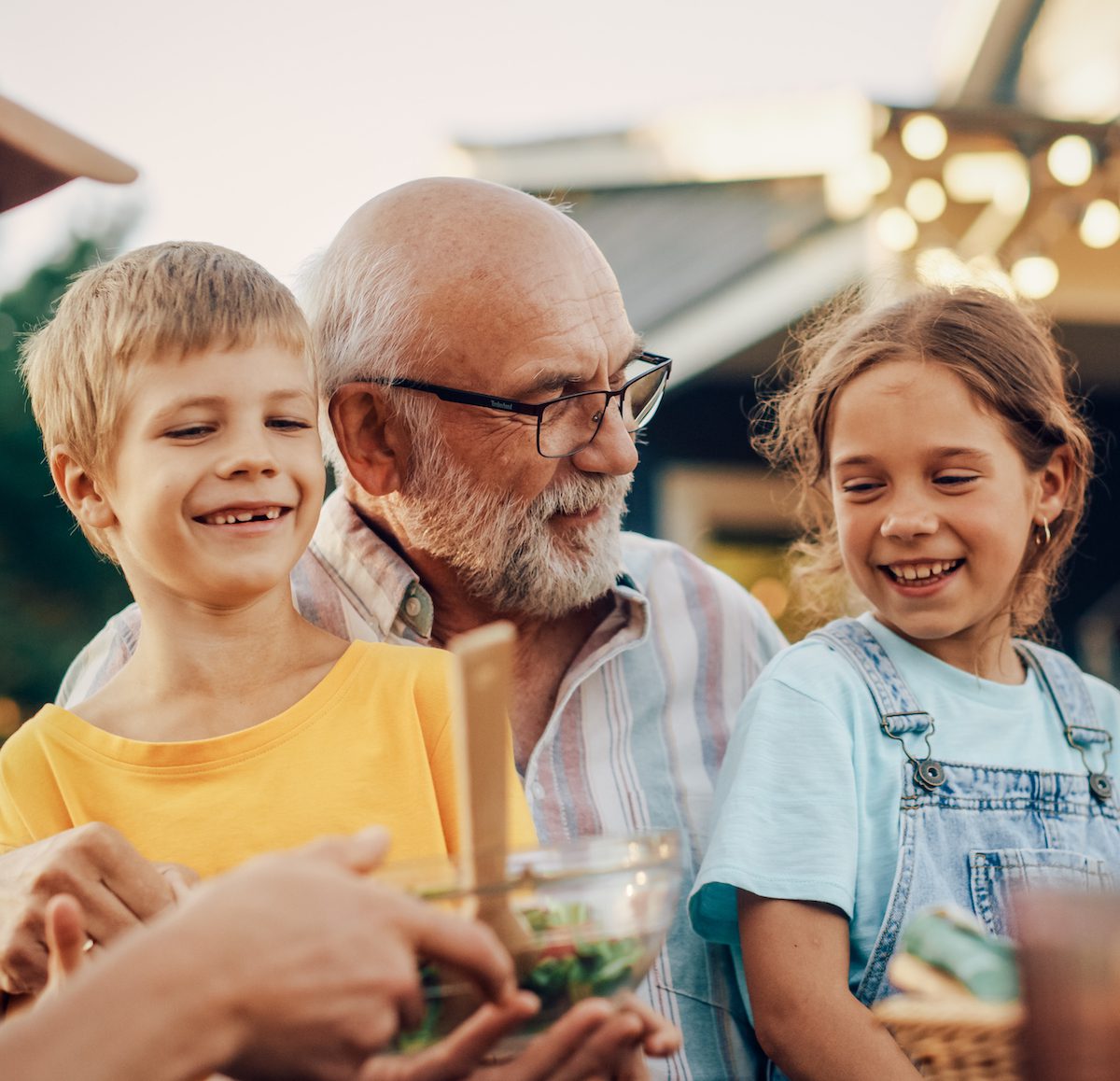 Happy Senior Grandfather Talking and Having Fun with His Grandchildren, Holding Them on Lap at a Outdoors Dinner with Food and Drinks. Adults at a Garden Party Together with Kids.