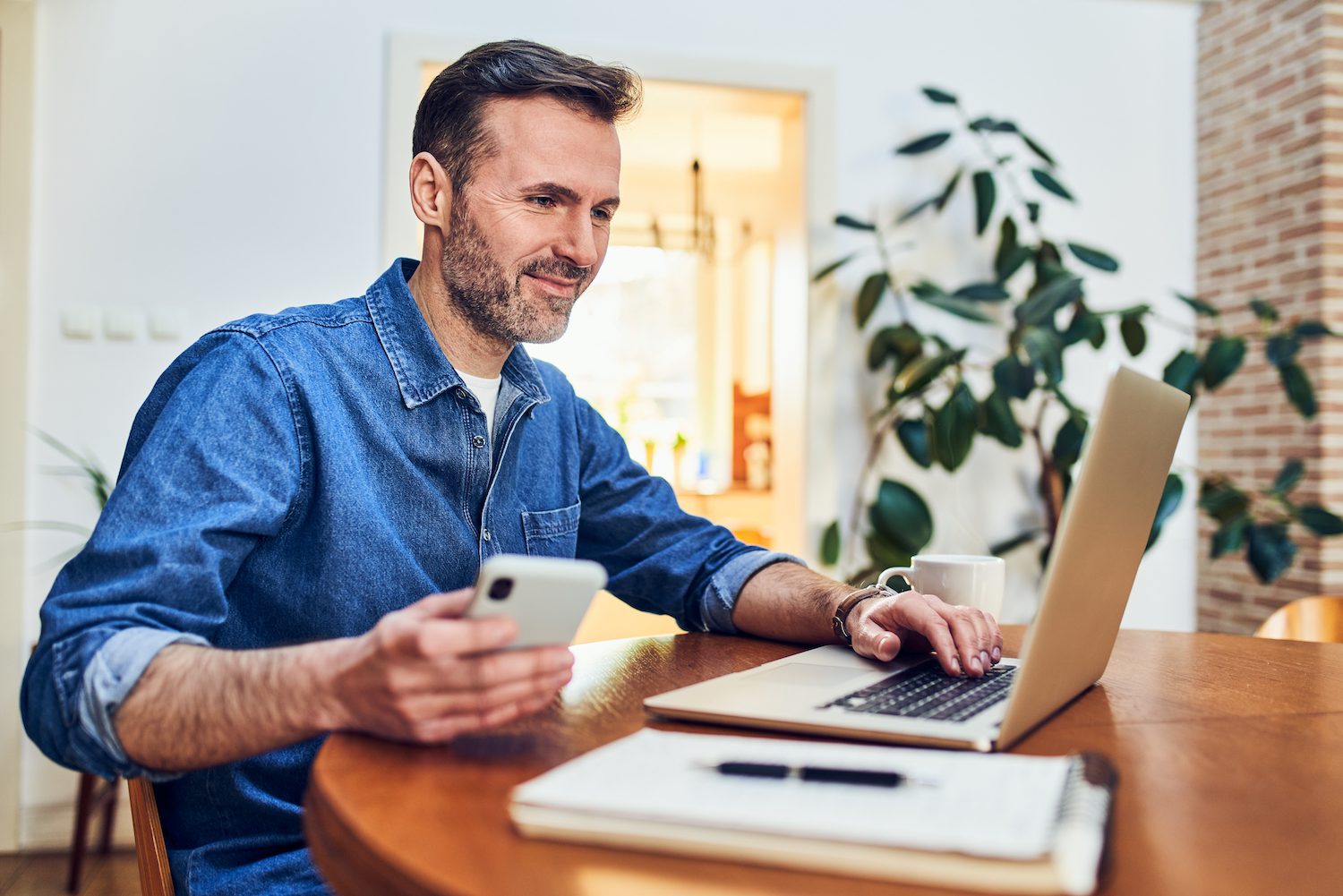 Smiling adult man using laptop and phone while freelancing at home